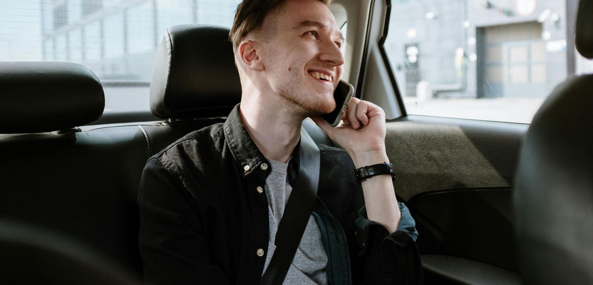 Smiling young man enjoying a phone call while seated in the back seat of a car during a daytime ride.